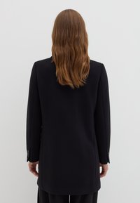 Manteau noir à la coupe ajustée, coupe droite, manches longues et poignets boutonnés, avec une texture lisse et un design minimaliste.