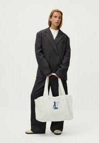 Baltas kanvas rankinės krepšys su mėlynomis siuvinėtomis raidėmis "L" ir "O". Turi du tvirtus rankenėles ir paprastą stačiakampio formą.