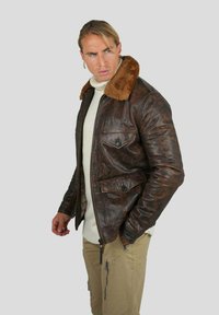 Veste en cuir marron avec un col en fausse fourrure, fermeture éclair à l'avant, deux poches poitrine et une finition texturée, portée sur un pull crème.