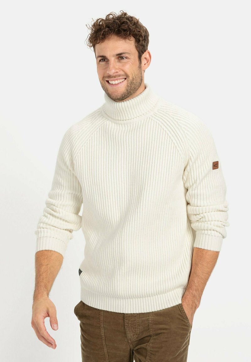 Weißer gerippter Rollkragenpullover mit hochgerolltem Kragen, langen Ärmeln und einem Lederpatch am linken Arm, kombiniert mit braunen Cordhosen.
