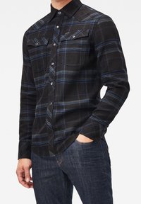 Camicia a quadri nera con accenti blu, maniche lunghe, colletto appuntito, due tasche sul petto e chiusure con bottoni metallici. Indossata con jeans scuri.
