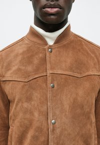 Detailný záber na osobu v hnedom zamšovom kabáte s gombíkmi, ktorý má na sebe biely rolák, zobrazuje textúru kabátu a kovové gombíky.