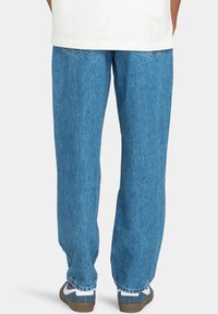 Vaqueros de mezclilla azul claro con corte relajado, con bolsillos traseros y una textura ligeramente desgastada. Se llevan con zapatillas azules y blancas.