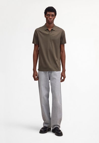 Homme debout, portant un polo à manches courtes vert olive foncé, un jean gris clair et des mocassins noirs, sur un fond blanc uni.