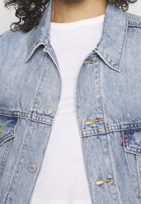 Světle modrá džínová bunda s límcem, měděným prošíváním a stříbrnými kovovými doplňky. Noste ji přes obyčejné bílé tričko.