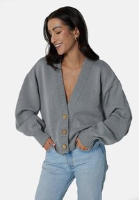 Szary sweter z dzianiny, z głębokim dekoltem w kształcie litery V i oversize'owymi rękawami. Pięć drewnianych guzików z przodu, ściągacze na mankietach i luźny krój.