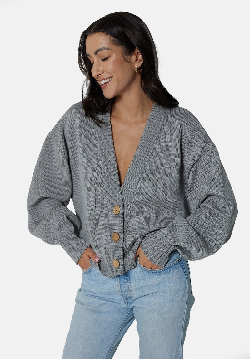 Szary sweter z dzianiny, z głębokim dekoltem w kształcie litery V i oversize'owymi rękawami. Pięć drewnianych guzików z przodu, ściągacze na mankietach i luźny krój.
