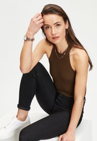 Brauner gerippter Tanktop, schwarze Jeans, weiße Sneakers. Accessoires mit einer silbernen Uhr und geschichteten Halsketten. Neutrale Hintergrund.