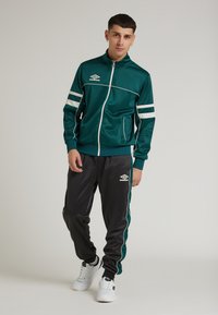 Grön sportjacka med vita ränder, dragkedjestängning och fickor, kombinerad med svarta byxor med gröna detaljer. Vita sneakers fulländar outfiten.