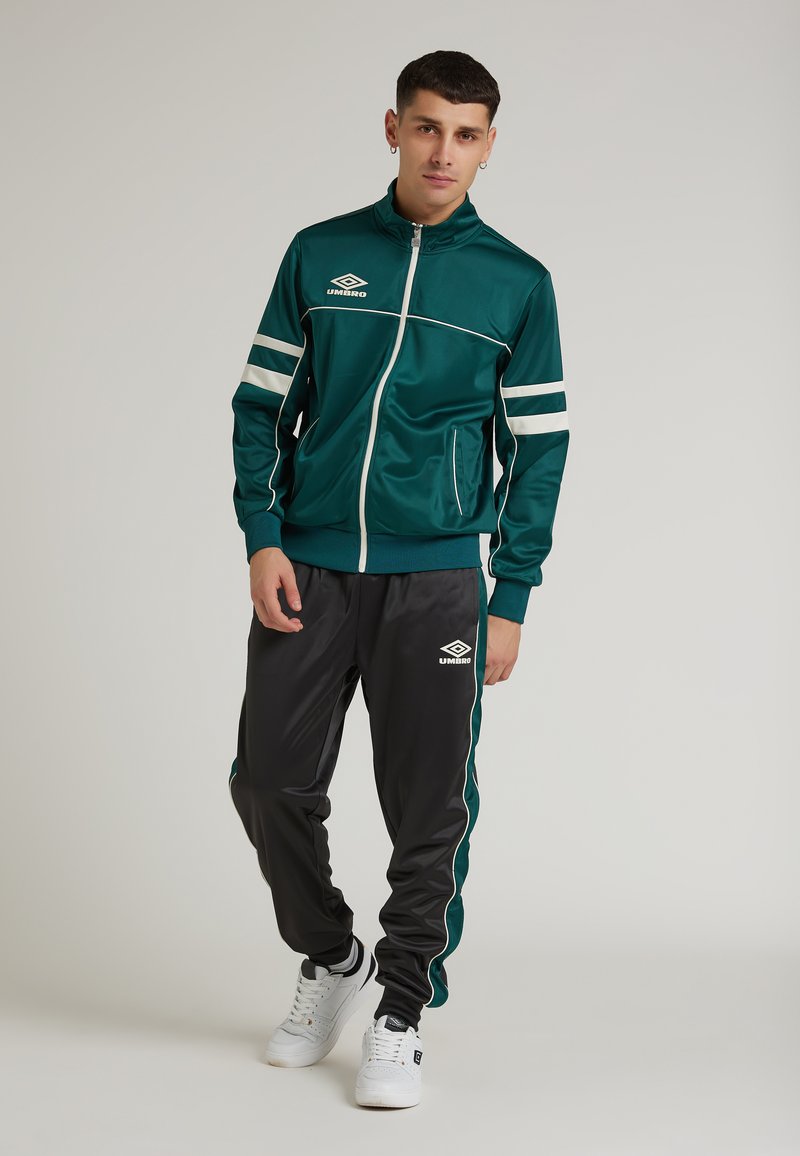 Grön sportjacka med vita ränder, dragkedjestängning och fickor, kombinerad med svarta byxor med gröna detaljer. Vita sneakers fulländar outfiten.