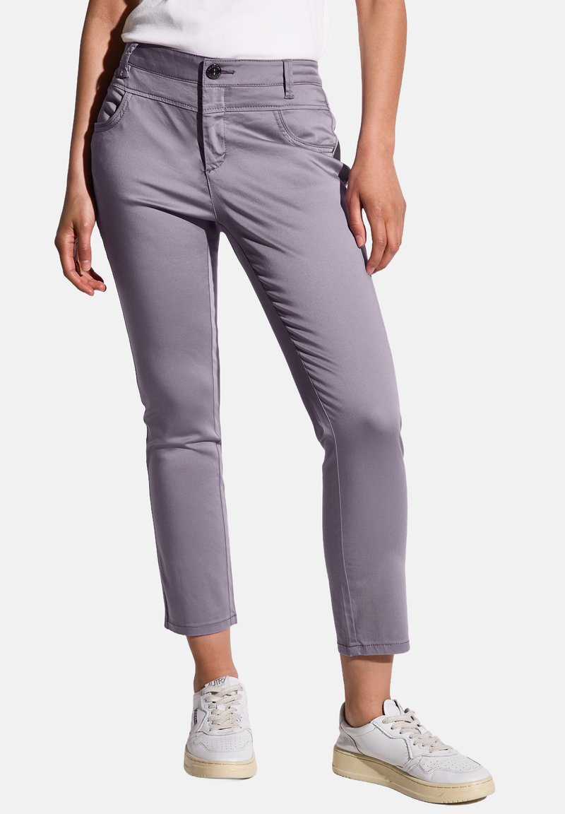 Chinos in lavanda con design aderente, chiusura con un bottone e tasche laterali; abbinati a sneakers bianche. Tessuto liscio e leggero.