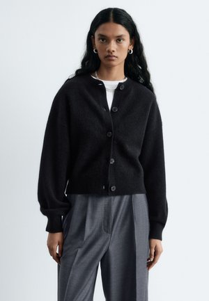 Femme portant un cardigan noir boutonné sur un haut blanc et un pantalon gris ample, avec de longs cheveux noirs et des boucles d'oreilles en argent en forme d'anneau.