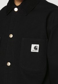 Musta corduroy-takki, jossa on kultaiset napit, rintatasku, jossa on valkoinen Carhartt-logo-patch ja klassinen kaulukuosi.