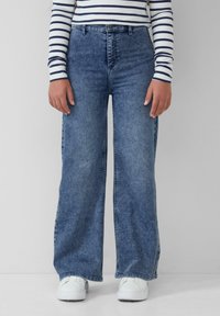 Džínsy so širokými nohavicami v stredne modrej džeme, s vysokým pásom, klasickými vreckami a viditeľným stehom. Skombinované s bielymi teniskami.