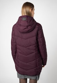 Vrouw met de rug naar de camera, gekleed in een lange, gewatteerde donkerpaarse hoodie-jack en een grijze gebreide rok, tegen een effen achtergrond.