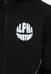 Black zip-up hoodie featuring a prominent white circular logo with the text "ALPHA INDUSTRIES." Material appears soft with a smooth texture.