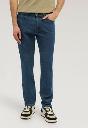 Man wearing straight-fit blue jeans and black-and-white sneakers, standing with one hand in pocket on neutral background.