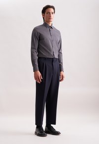 Herenoutfit met een getailleerd, gemusterd overhemd in marineblauw met witte accenten, gecombineerd met hoogmixte, donkergekleurde broeken en zwarte veterschoenen.