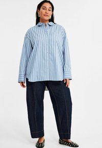 Helles blau gestreiftes Oversized-Hemd mit Knopfverschluss, langen Ärmeln und einer Brusttasche, kombiniert mit dunkelblauen weiten Jeans mit Kontrastnähten.