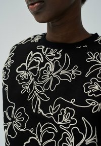 Pull noir avec broderie florale blanche, col rond et tissu texturé. Gros plan mettant en avant les détails du design et les motifs de couture.