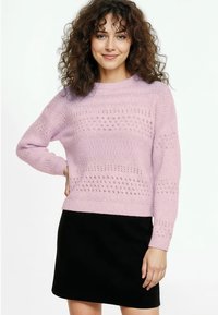 Világos rózsaszín kötött pulóver kerek nyakkivágással, mintás textúrával és hosszú ujjakkal, fekete szoknyával párosítva.