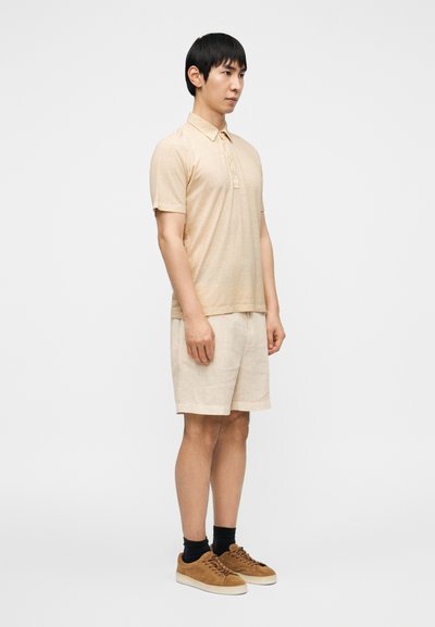 Μπεζ πόλο πουκάμισο με κοντά μανίκια και τρία κουμπιά, συνδυασμένο με ανοιχτό μπεζ σορτς και καφέ σουέντ αθλητικά παπούτσια. Φαίνονται μαύρες κάλτσες.