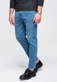 Jean en denim bleu clair à coupe droite, avec des détails subtils de délavage, associé à des chaussures en cuir noir et un pull foncé.