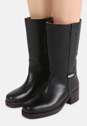 Crne kožne čizme srednje dužine do lista s niskom blok petom, bočnim patentnim zatvaračem, suptilnim detaljima šavova i malom metalnom oznakom brenda na vanjskoj strani.