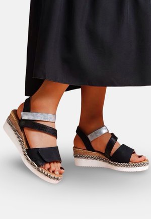 Pieds portant des sandales compensées noires et argentées avec des semelles à motifs, associées à une jupe noire arrivant mi-mollet.