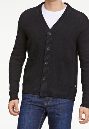 Mann trägt eine schwarze, gerippte Strickjacke mit Knöpfen und Fronttaschen über einem weißen Hemd und blauen Jeans.