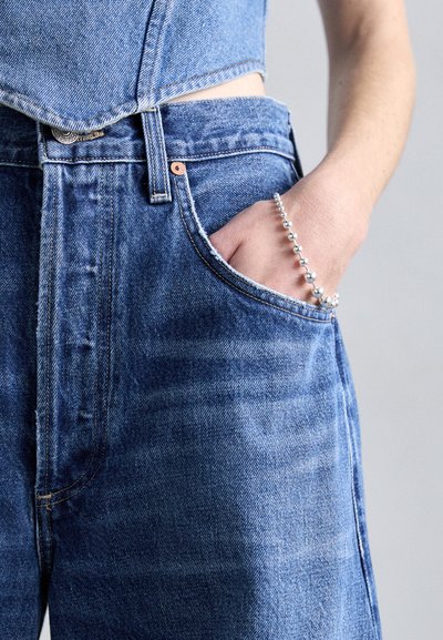 Jean en denim bleu taille haute avec une texture délavée, présentant une poche avant et une main ornée d'un bracelet en perles argentées.