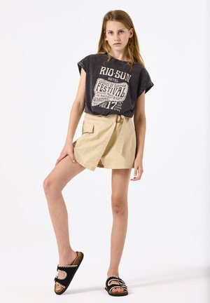 Grijze graphic t-shirt met korte mouwen, gecombineerd met beige wikkelshorts met een zijzak en een riem. Zwarte sandalen met witte accenten.