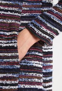 Tejido texturizado con rayas verticales en marino, blanco, rosa y azul. Presenta una mano en un bolsillo lateral y bordes de mangas deshilachados.