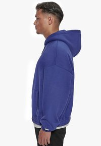 Haut à capuche violet avec une grande capuche, une poche kangourou et des poignets et un ourlet côtelés. Fait en tissu doux avec une finition légèrement texturée.