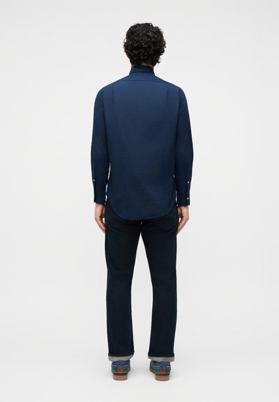 Göndör hajú férfi hosszú ujjú tengerészkék ingben, sötét, felhajtott szárú farmerben és kék cipőben, háttal áll egy egyszerű háttér előtt.