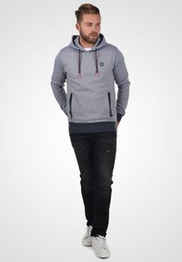 Grauer Kapuzenpullover mit strukturiertem Finish, Fronttasche und verstellbaren Kordeln. Kombiniert mit schwarzen, zerrissenen Jeans und weißen Sneakern.