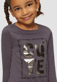 Langärmliges lila Oberteil mit großen metallisch goldenen und schwarzen Buchstaben, die "CUTE" sagen, während die Phrase "is my magic" in kleinerer Schrift wiederholt wird.