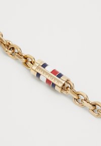Kuldne käevõru silindrilise lukuga, millel on punane, valge ja sinine triip ning sellele on graveeritud "HILFIGER."