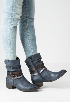 Bottes en cuir bleu mi-mollet avec des sangles marron décoratives et des fermetures éclair, portées avec un jean skinny bleu clair.