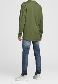 Man med olivgrön långärmad skjorta, blå jeans och vita sneakers, står vänd bort från kameran mot en enfärgad bakgrund.