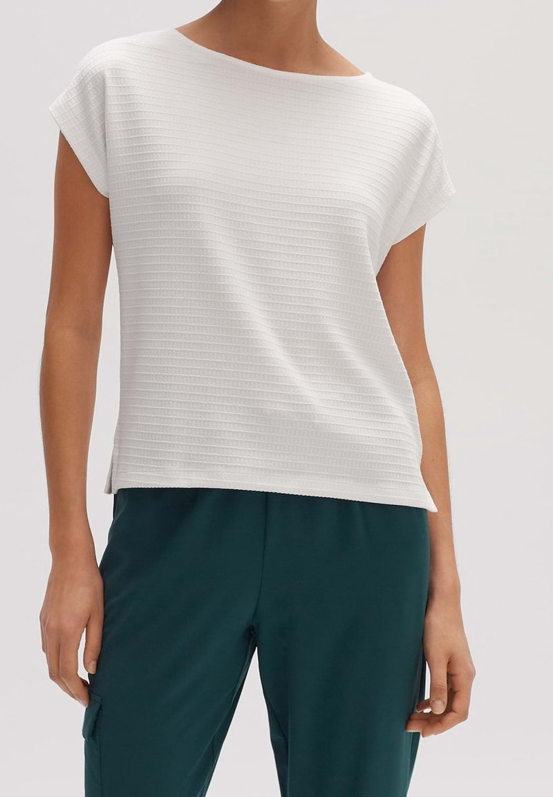 Personne portant un haut blanc texturé à manches courtes et un pantalon vert foncé avec poches latérales, montré du cou aux hanches.