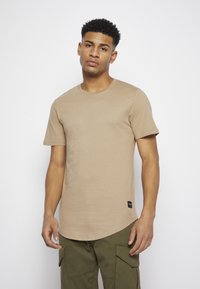 T-shirt de manga curta bege em tecido suave com decote redondo, apresentando uma bainha curva e uma pequena etiqueta da marca preta na parte inferior.