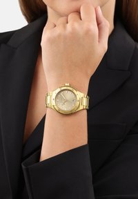 Goldene Armbanduhr mit rundem Ziffernblatt, beigem Zifferblatt, Diamantmarkierungen und strukturiertem Gehäuse. Die Glieder sind poliert und passen zu einem schwarzen Blazerärmel.