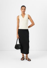 Ärmelloses cremefarbenes Oberteil mit V-Ausschnitt und Raffung, kombiniert mit einem langen schwarzen Rock. Das Model trägt eine kleine schwarze Handtasche und schwarze Riemchensandalen.