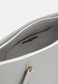 Šedá texturovaná kožená taška přes rameno se zipovým uzávěrem, s vnitřkem z světle šedé látky a malou vnitřní kapsou označenou "ANNA FIELD."