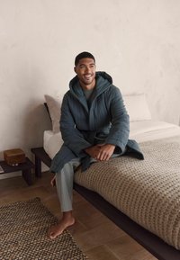 Gestepptes türkisfarbenes Badekleid mit Kapuze, getragen über einem grauen Oberteil und gestreiften Schlafanzughosen. Sitzt auf einem Bett mit einer strukturierten Decke und einem Holznachttisch.