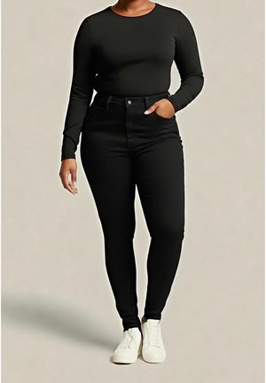 Person iført sort langærmet skjorte og højtaljede sorte skinny jeans med hvide sneakers, stående med den ene hånd i lommen.