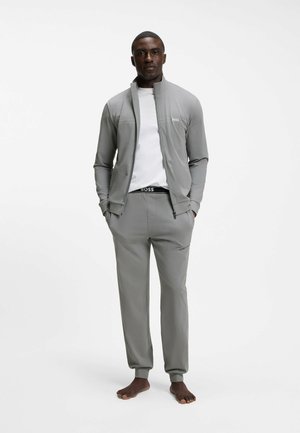 Man standing barefoot wearing gray zip-up jacket, matching pants, and white T-shirt, hands in pockets, against plain white background.