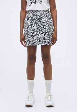 Asmuo, dėvintis baltą sijoną su leopardo raštu, baltą grafišką marškinėlį, baltas kojines ir baltus sportbačius, stovintis prieš paprastą foną.