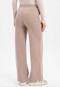 Osoba w beżowych spodniach z szerokimi nogawkami, kremowej, prążkowanej bluzce z długim rękawem i szarych sneakersach Adidas stoi na białym tle.